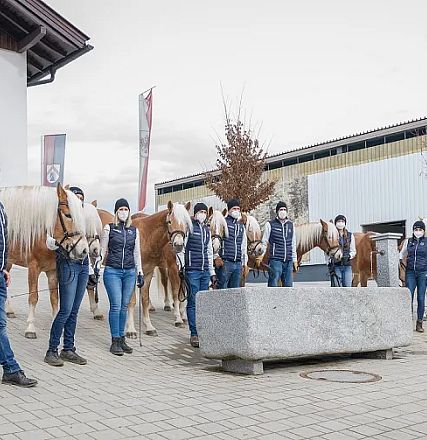 Eine Gruppe von Menschen steht mit Pferden vor einem Gebäude. Alle tragen identische Kleidung und Masken, unter einem bewölkten Himmel.
