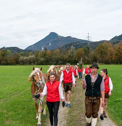 Eine Gruppe von Menschen in traditioneller Kleidung führt Pferde über einen Feldweg, umgeben von grünen Wiesen und Bergen im Hintergrund.