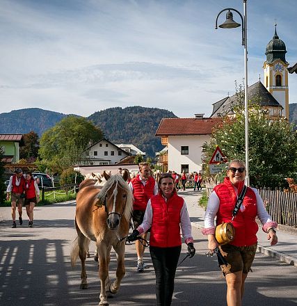 Eine Gruppe von Menschen in traditionellen Trachten führt ein Pferd durch ein malerisches alpenländisches Dorf mit bunten Häusern und einer Kirchturmspitze.