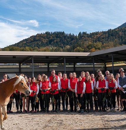 Gruppe von Menschen in roter Kleidung steht vor einem Pferdegehege mit zwei Pferden. Im Hintergrund sind Berge und ein blauer Himmel zu sehen.