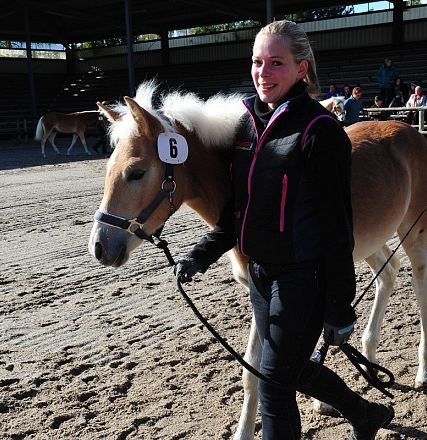 Donna in un'arena guida un cavallo con il numero sei. Indossa abbigliamento scuro e sorride, mentre il pubblico osserva dagli spalti.