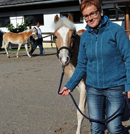 Donna sorridente con occhiali conduce un giovane cavallo in un'area all'aperto. Altri cavalli e persone si vedono sullo sfondo davanti a un edificio.