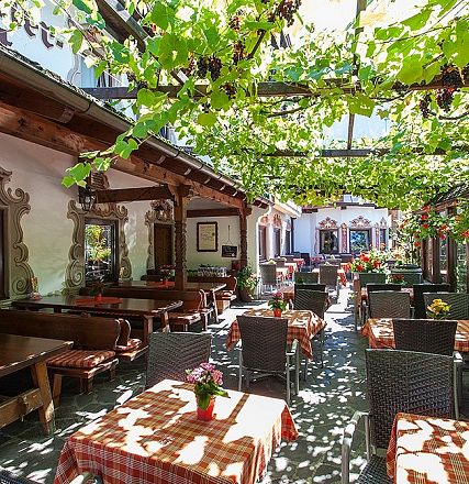 Ein gemütlicher Biergarten mit Holzmöbeln und rot-weiß karierten Tischdecken, von einem grünen Blätterdach beschattet. Im Hintergrund traditionelle Architektur.