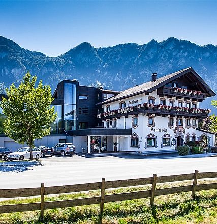 Ein modernes Hotelgebäude neben einem traditionellen Alpenhaus, beides vor einer Bergkulisse. Die Mischung aus alt und neu ist harmonisch angeordnet.