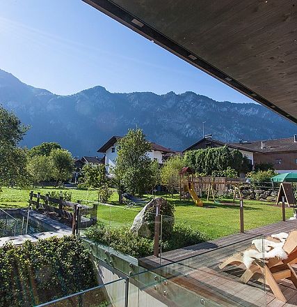 Ein sonniger Garten mit Bergblick. Im Vordergrund eine Terrasse mit Holzmöbeln, im Hintergrund grüne Bäume und Häuser vor beeindruckenden Bergen.