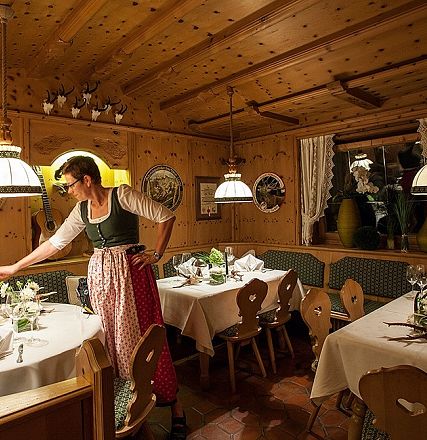 Gemütliches bayerisches Restaurant mit Holzwänden und traditioneller Dekoration. Eine Frau in Dirndl deckt die Tische mit weißen Tischdecken und Blumen.