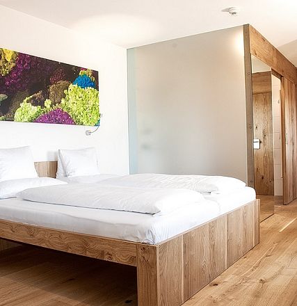 Modernes Hotelzimmer mit Holzbett und Parkettboden. An der Wand hängt ein buntes Gemälde. Das Zimmer wirkt hell und einladend mit schlichtem Design.