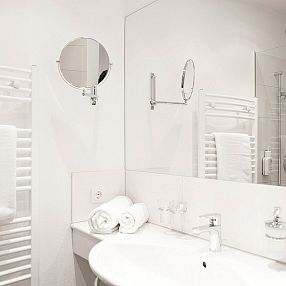 Bagno moderno con pareti bianche, lavabo, specchio grande, asciugamani arrotolati sul lavandino e una doccia in vetro con soffione.