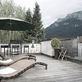 Terrazza panoramica con sedie a sdraio e ombrellone, circondata da alberi e montagne in lontananza. Atmosfera rilassante e paesaggio naturale.