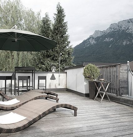 Holzterrasse mit Liegestühlen, großem Sonnenschirm und Pflanzen; im Hintergrund Berglandschaft und bewölkter Himmel. Ruhige, entspannende Atmosphäre.
