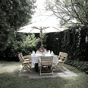 Giardino ombreggiato con tavolo apparecchiato, circondato da sedie in legno e piante verdi. Un grande ombrellone bianco offre riparo dal sole.