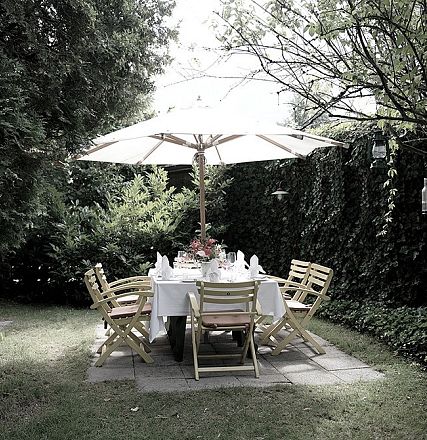 Gemütlicher Garten mit einem gedeckten Tisch unter einem Sonnenschirm, umgeben von üppigen grünen Pflanzen und Holzstühlen. Perfekt für ein Sommeressen.