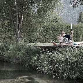 Due persone sedute su una piattaforma in legno vicino a un ruscello, circondate da alberi e prati verdi. Sembrano rilassarsi e godersi la natura.