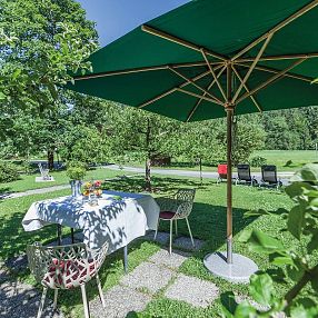 Ein grüner Garten mit einem Tisch unter einem großen Sonnenschirm. Der Tisch ist mit einer weißen Tischdecke gedeckt. Im Hintergrund sind Bäume und ein Wiese.