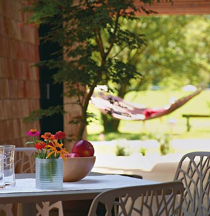Ein entspannter Gartenbereich mit einem Tisch, auf dem Blumen und Obst stehen. Im Hintergrund hängt eine Hängematte zwischen zwei Bäumen.