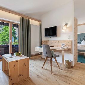 Modernes, helles Hotelzimmer mit Holzmöbeln, Schreibtisch, Stuhl, Bett und Balkon mit Blick auf grüne Bäume. Der Raum wirkt einladend und gemütlich.