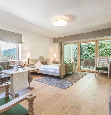 Helles Zimmer mit Holzboden, stilvollen Möbeln und großen Fenstern. Ein Einzelbett, ein Tisch mit Stühlen und ein Zugang zu einem Balkon mit Blick ins Grüne.