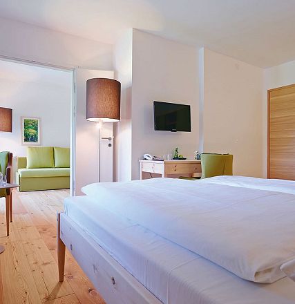 Helles, modernes Hotelzimmer mit einem großen Bett, Holzmöbeln und einem Flachbildfernseher. Im Hintergrund sind eine Sitzecke und helle Wände zu sehen.