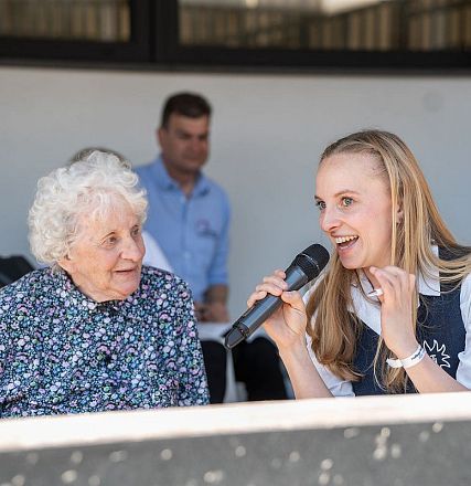 An elderly woman with curly white hair and a patterned blouse sits beside a young woman holding a microphone, speaking at an outdoor event.