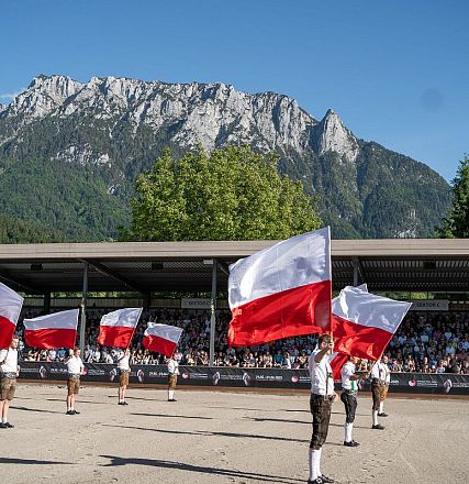 Mehrere uniformierte Personen schwenken rot-weiße Fahnen auf einem Platz vor einer beeindruckenden Berglandschaft und einem Publikum auf Tribünen.