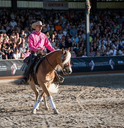 A rider in a bright pink shirt and cowboy hat performs dressage on a brown horse in an arena, with a large crowd watching under a shaded area.