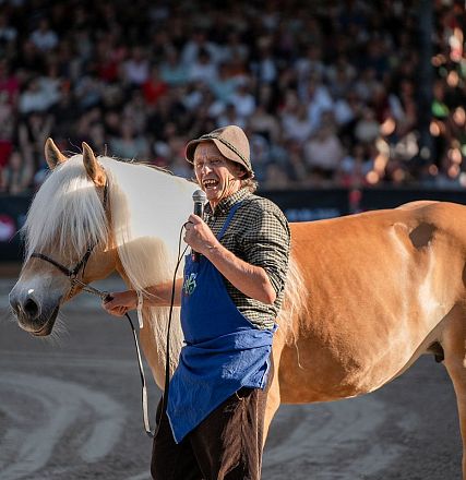 Un uomo con un cappello parla al microfono accanto a un cavallo biondo. Sullo sfondo, una folla assiste all'evento all'aperto in un'arena.