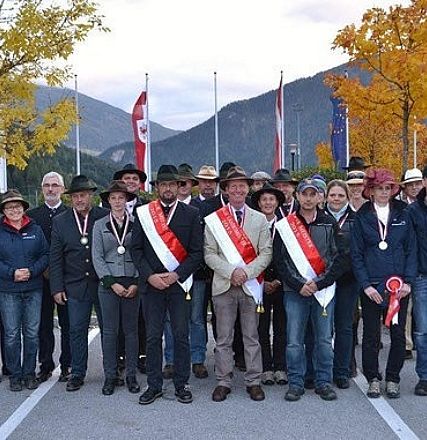 Gruppo di persone in abiti tradizionali, alcune con medaglie, posano all'aperto con montagne e alberi autunnali sullo sfondo.