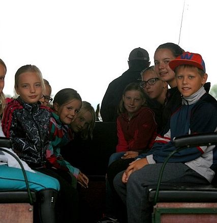 Un gruppo di bambini sorridenti è seduto su una carrozza trainata da cavalli. Indossano abiti colorati e sembrano divertirsi durante una giornata all'aperto.