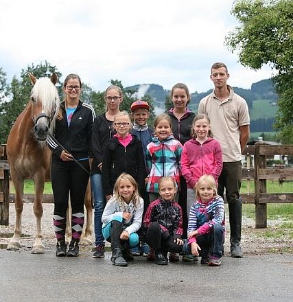 Gruppo di persone in piedi all'aperto davanti a un cavallo marrone chiaro e una recinzione di legno. Sullo sfondo ci sono alberi e colline verdi.