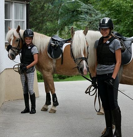 Due amazzoni sorridenti in abbigliamento da equitazione accanto ai loro cavalli marroni e bianchi, pronte per una passeggiata in una giornata serena.