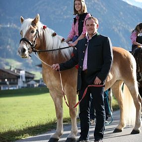 Un cavallo biondo con una criniera decorata accanto a un uomo in abiti tradizionali tirolesi. Sullo sfondo, paesaggio di montagne e casette.