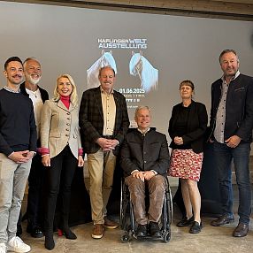 Un gruppo di persone in posa davanti a uno schermo, abbigliamento formale. Sullo schermo, un logo e una data. L'ambiente sembra essere una sala conferenze.