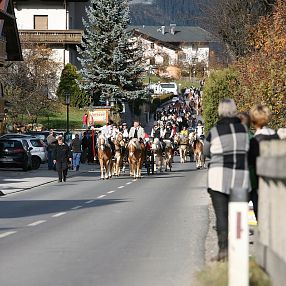 Una strada di campagna con persone accanto ai carri trainati da cavalli durante una sfilata. Alberi e case decorano il paesaggio autunnale.
