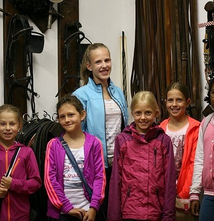 Gruppo di sei ragazze con giacche colorate, sorridenti in una stanza con attrezzature equestri appese alle pareti, atmosfera amichevole e accogliente.