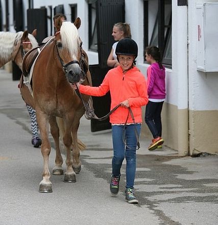 Una bambina in felpa arancione conduce un cavallo marrone chiaro con una criniera bianca lungo una strada acciottolata. Dietro di loro ci sono altre due persone.