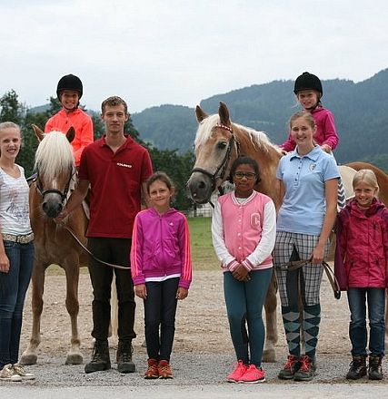 Un gruppo di bambini e adulti posano con due cavalli biondi in un ambiente rurale, con alberi e montagne sullo sfondo.