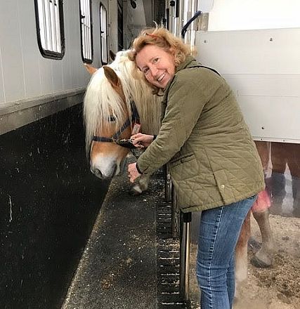 Frau streichelt ein helles Pferd in einem Stall, beide wirken entspannt und zufrieden. Die Frau trägt eine grüne Jacke und das Pferd hat eine lange Mähne.