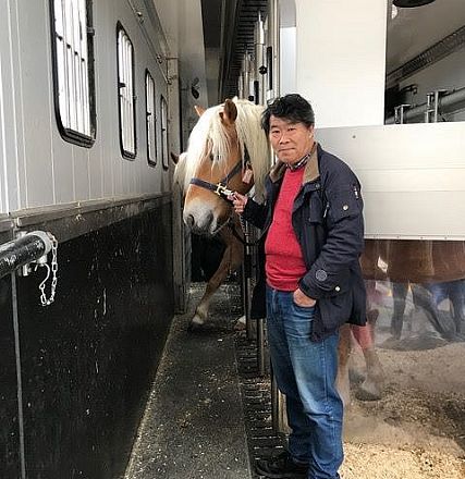 Mann steht in einem Pferdetransportanhänger neben einem Pferd mit heller Mähne. Er hält einen Führstrick und trägt eine blaue Jacke und Jeans.