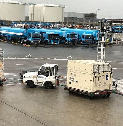 Ein Industrielagerplatz mit mehreren blauen Tanklastwagen und gestapelten Containern auf nassem Asphalt. Ein weißer Schlepper transportiert einen großen Container.