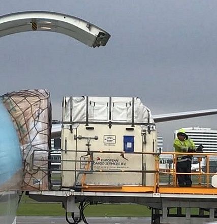 Flughafenpersonal lädt eine große weiße Frachtkiste in das geöffnete Heck eines Flugzeugs bei bewölktem Himmel. Die Szene spielt sich auf dem Rollfeld ab.