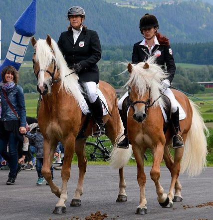 Two people riding horses on a paved road, surrounded by mountains and green fields. An inflatable bouncy structure is visible in the background.