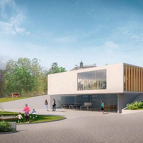 Moderne Gebäude mit großen Glasfenstern, umgeben von grüner Landschaft und Bäumen. Im Vordergrund Menschen beim Spaziergang und ein Kinderspielplatz.