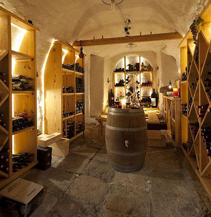 Ein stimmungsvoller Weinkeller mit gedämpfter Beleuchtung, gefüllten Weinregalen und einem Holzfass im Zentrum. Die Wände sind aus rustikalem Stein.