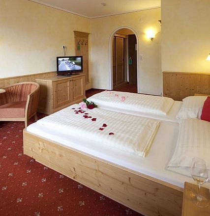 Gemütliches Hotelzimmer mit zwei Betten, dekoriert mit Rosenblättern. Holzmöbel und roter Teppichboden. Weinglas und TV schaffen eine einladende Atmosphäre.