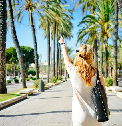 Eine Frau mit blonden Haaren, Kopfhörern und einer großen Tasche geht auf einer von Palmen gesäumten, sonnigen Promenade und zeigt in die Ferne.