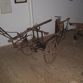 Altes, handbetriebenes landwirtschaftliches Gerät mit Metallrädern und hölzernen Griffen auf Holzboden, möglicherweise ein Pflug oder eine Egge.