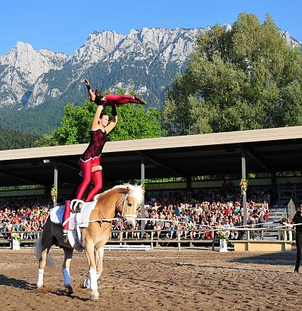 Ein Artist führt eine Akrobatiknummer auf einem weißen Pferd vor einer Tribüne mit Publikum auf. Im Hintergrund sind bewaldete Berge sichtbar.