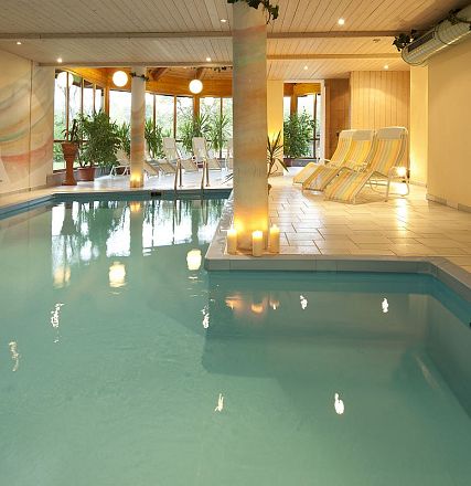 Ein ruhiger Innenpool mit blauem Wasser, umgeben von Kerzenlicht und Pflanzen. Im Hintergrund stehen Liegestühle und große Fenster bieten Tageslicht.