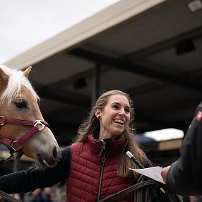 Eine lächelnde Frau in roter Weste steht neben einem hellbraunen Pferd mit blonder Mähne. Sie unterhält sich mit einer Person, die Notizen hält.