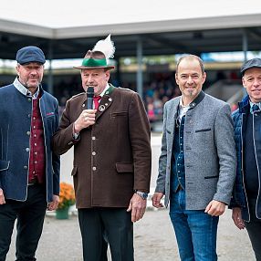 Vier Männer stehen auf einem öffentlichen Platz, sie tragen traditionelle bayerische und moderne Kleidung. Einer hält ein Mikrofon. Im Hintergrund sind Zuschauer zu sehen.